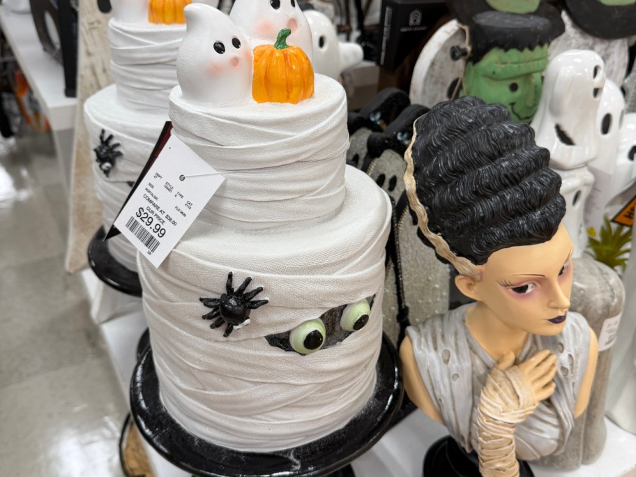 A white mummy-wrapped tiered figurine topped with ghosts and a pumpkin, accompanied by a Frankenstein&rsquo;s bride bust.