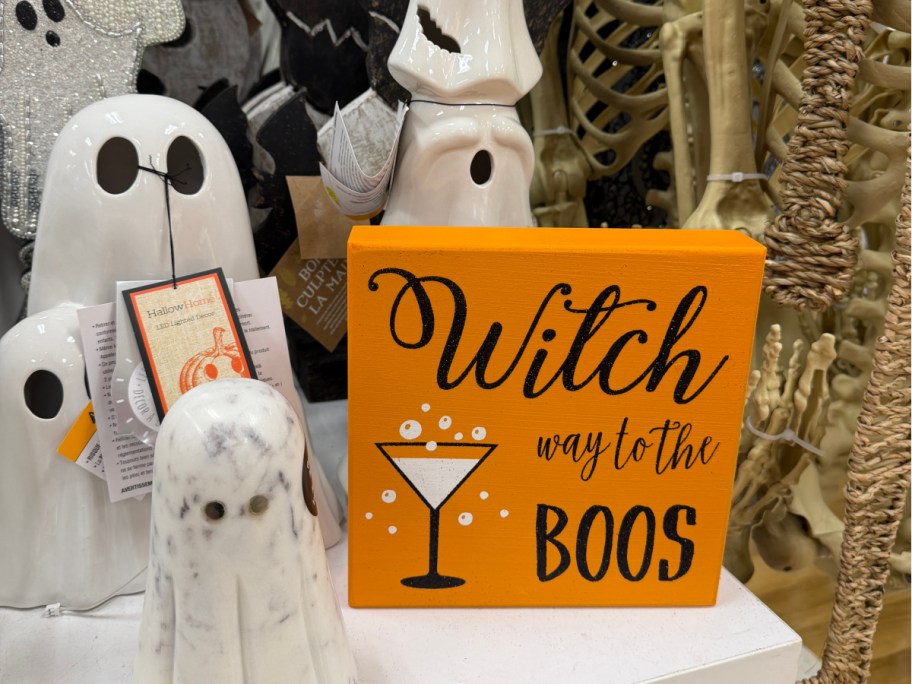 An orange sign that reads "Witch way to the BOOS" with a martini glass graphic, displayed alongside white ghost figurines and Halloween decor.