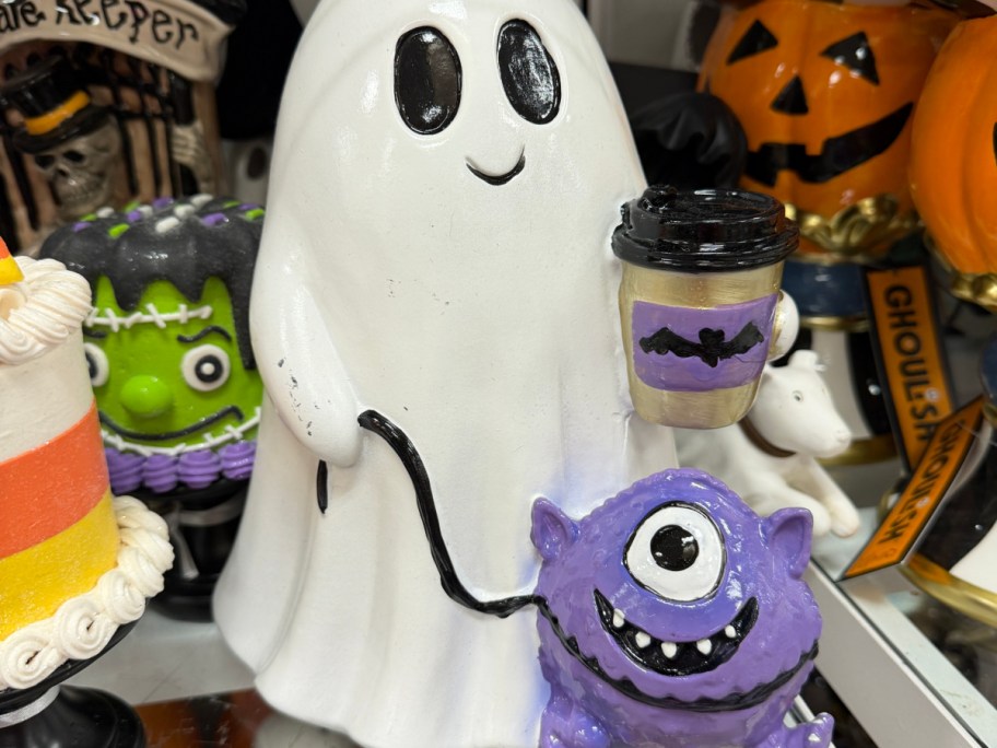 A white ghost figurine holding a coffee cup and walking a small purple one-eyed monster on a leash, surrounded by other Halloween decor.