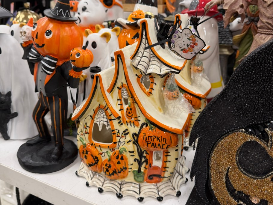 A ceramic Halloween "Pumpkin Palace" house figurine decorated with painted pumpkins and spiderwebs, surrounded by other holiday decor on a store shelf.