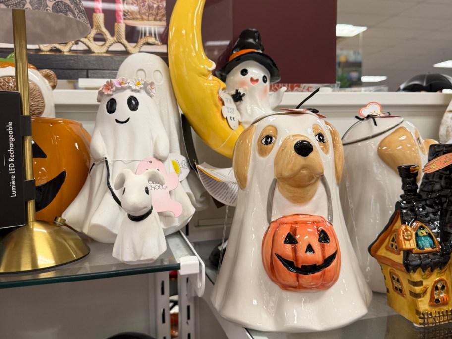 A ceramic ghost figurine dressed as a dog holding a pumpkin candy bucket, surrounded by other Halloween decor on a shelf.