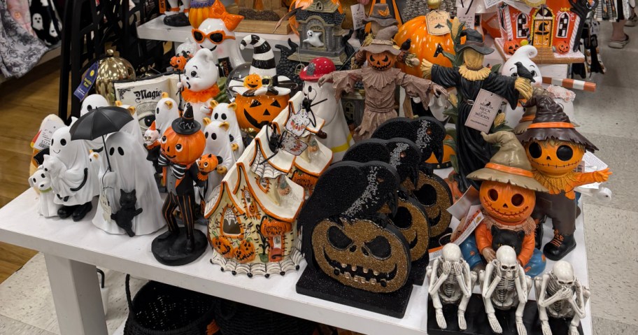 A store display table stocked with various Halloween decor items, including ghost figurines, pumpkin-headed figures, scarecrows, glittery jack-o'-lanterns, and skeleton figurines.
