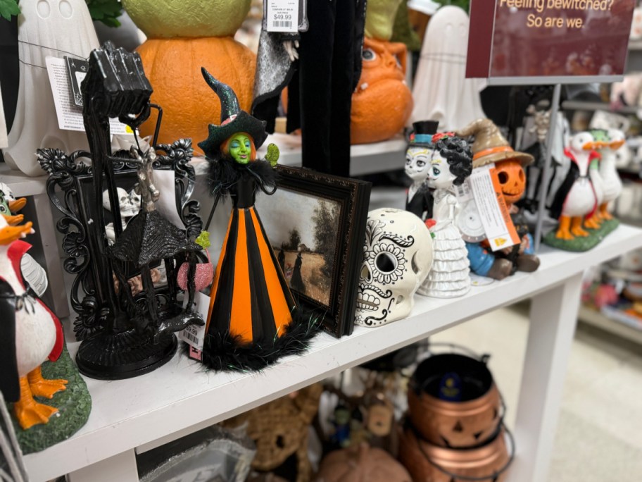A witch figurine with a green face and an orange and black striped dress displayed on a store shelf among other Halloween decorations.