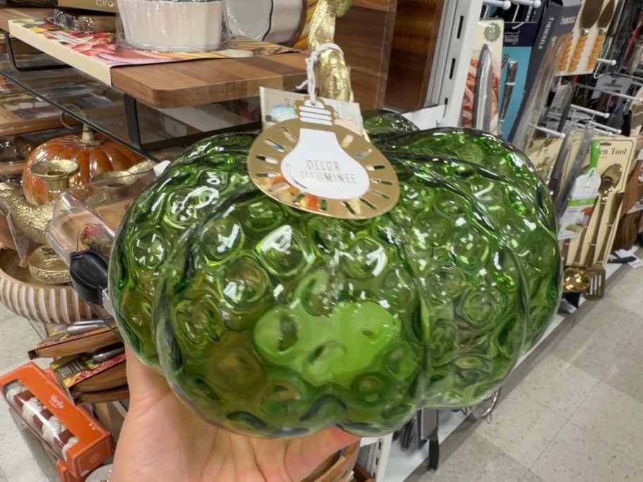A green glass pumpkin figurine with a textured, bumpy surface and a gold stem being held in a store.