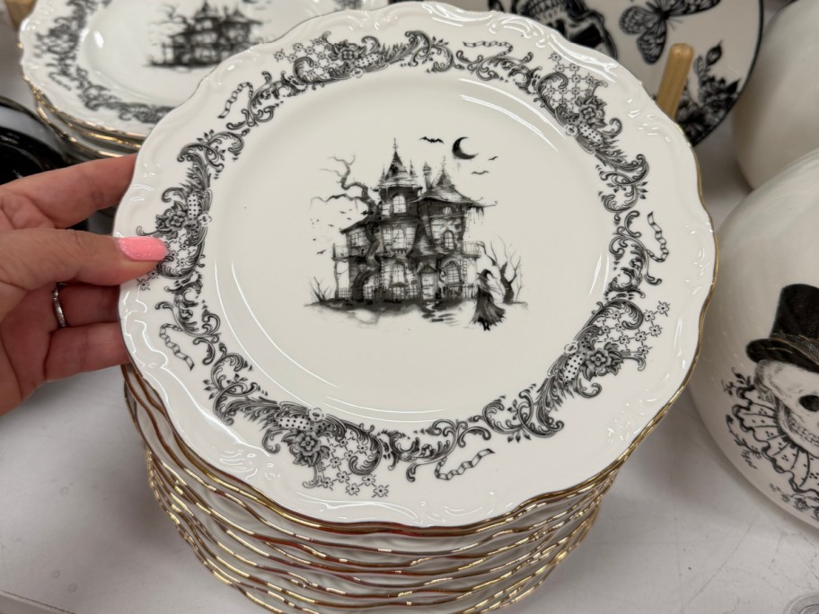 A stack of white ceramic plates featuring an ornate black floral rim, gold trim, and a central illustration of a haunted house.
