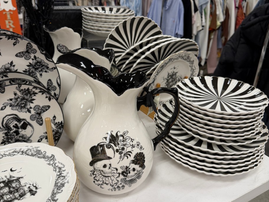 A white ceramic pitcher featuring a black-and-white sketch of two skeletons in top hats, displayed alongside black-and-white striped plates and floral tableware.