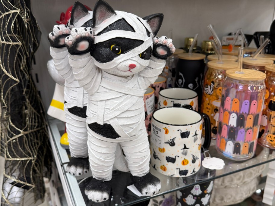 A black and white cat figurine wrapped like a mummy with its paws up, standing on a store shelf next to Halloween mugs.
