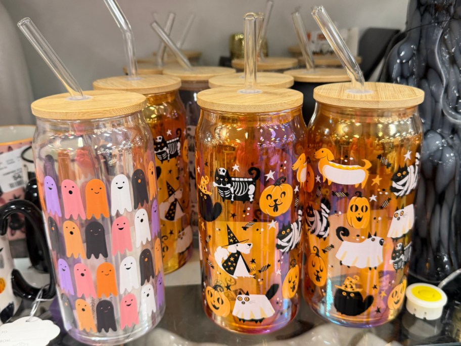 A collection of glass tumblers with bamboo lids and glass straws, featuring colorful Halloween designs of ghosts, cats, and pumpkins.