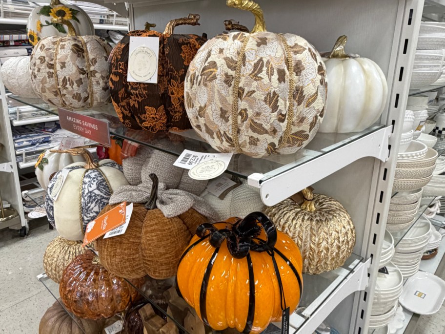A store shelf displaying a variety of decorative autumn pumpkins in different textures, colors, and patterns, including fabric, velvet, and metallic designs.