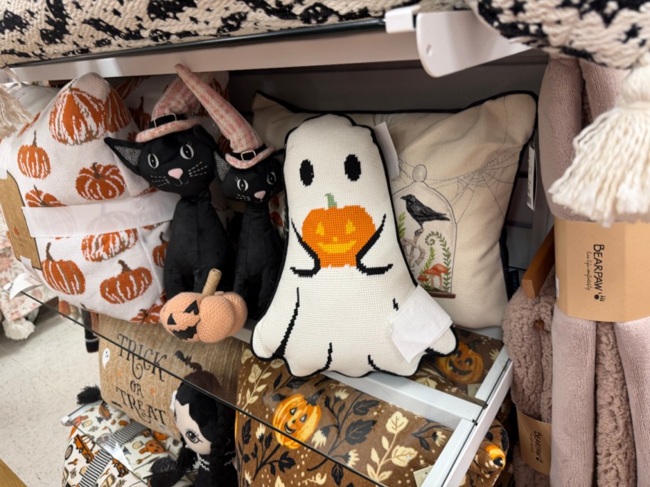 A ghost-shaped decorative pillow holding a pumpkin, displayed on a store shelf alongside black cat figurines and other Halloween pillows.