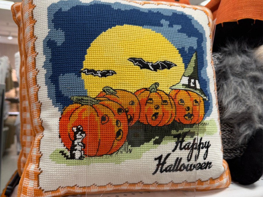 A decorative Halloween pillow with a stitched design of four jack-o'-lanterns, bats against a yellow moon, a cat, and the words "Happy Halloween."