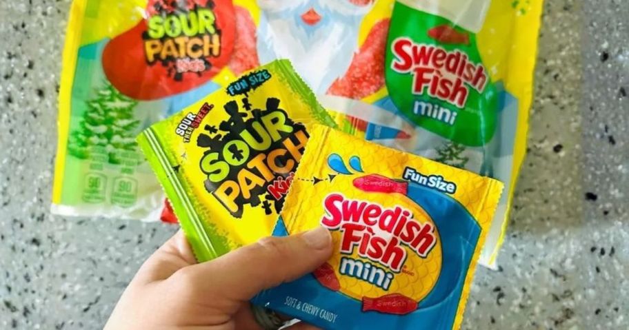 hand holding sour patch kids and swedish fish packs above bag on counter