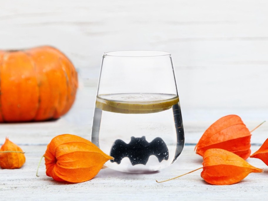 A clear stemless glass filled with water and a floating lemon slice, featuring a small black bat figure at the bottom, sitting on a white wooden surface surrounded by orange lantern pods and a pumpkin.