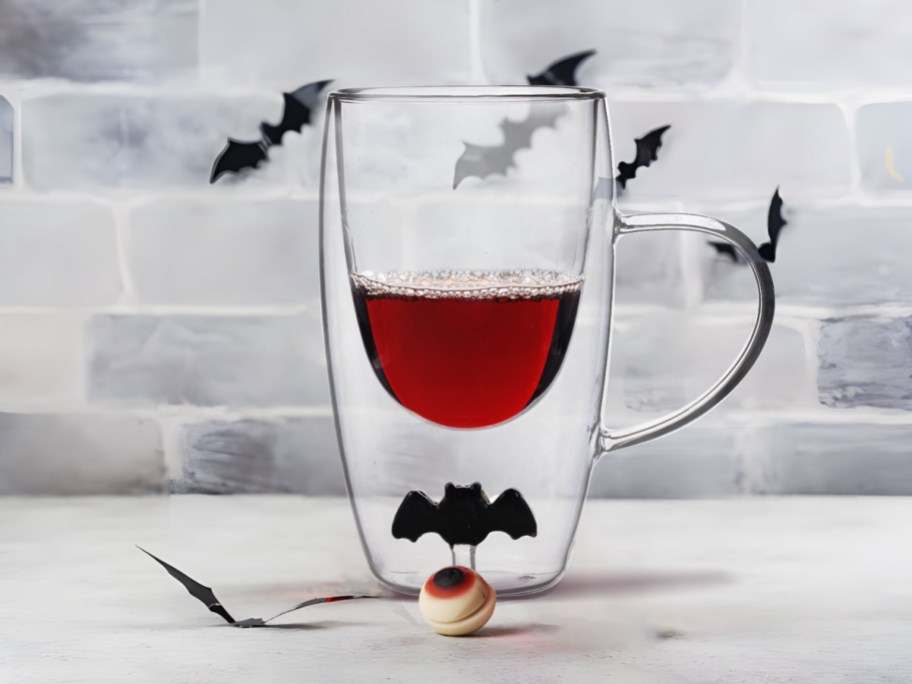 A double-walled clear glass mug partially filled with red liquid, featuring a small black bat figure inside the base of the glass, set against a white brick wall decorated with black bat silhouettes.