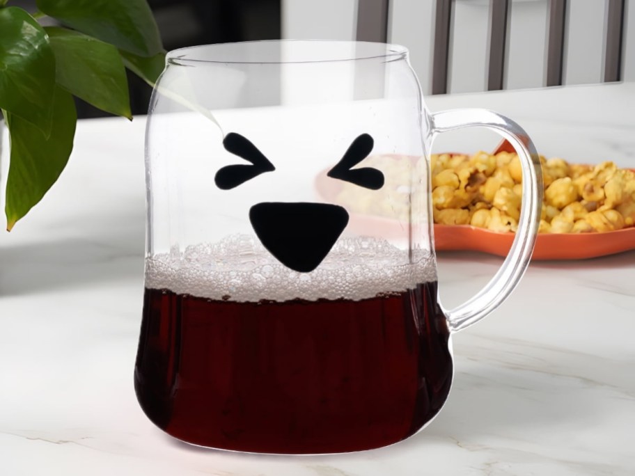 A clear glass mug featuring a printed black happy face, partially filled with a dark foamy beverage.