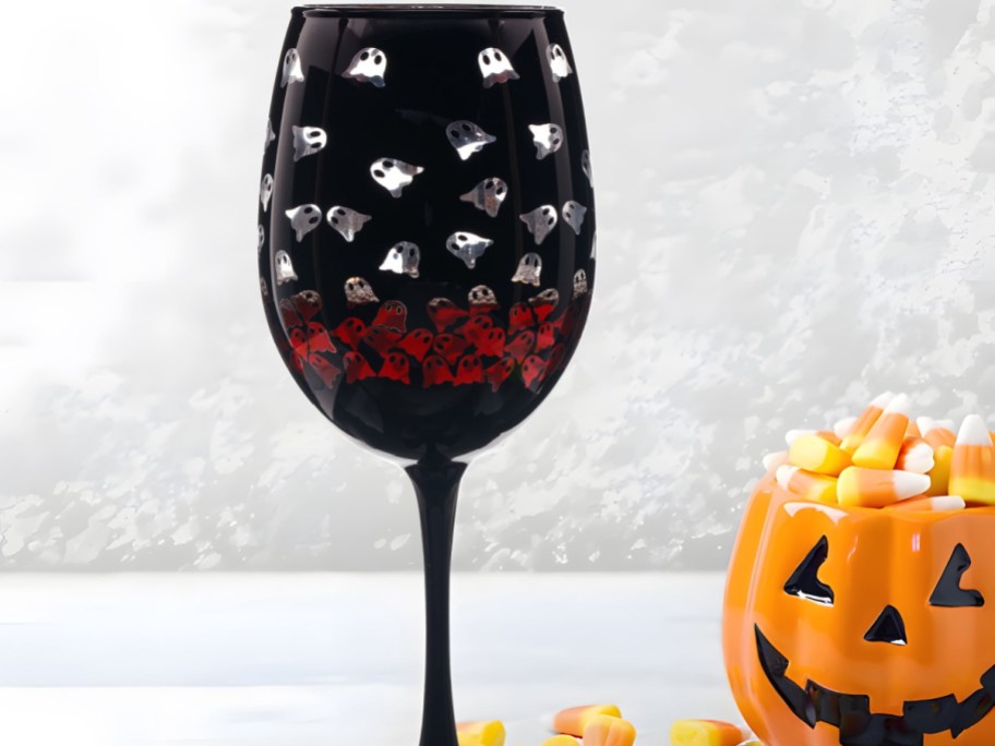 A black stemmed wine glass patterned with small silver and red ghosts, standing next to a ceramic jack-o'-lantern bowl filled with candy corn.