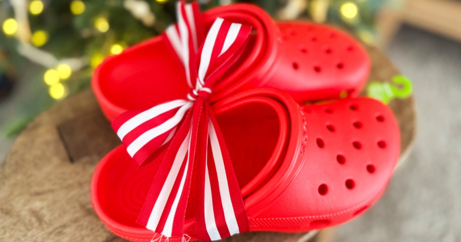 pair of red crocs clogs with red and white ribbon around them near christmas tree