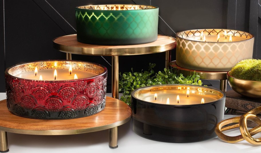 Four large, multi-wick luxury candles in decorative glass jars displayed on tiered stands.