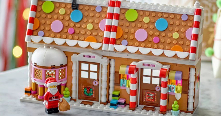 A completed LEGO gingerbread house decorated with colorful candy-like pieces on the roof, white scalloped frosting details, and candy cane columns, with a Santa Claus minifigure standing in front holding a brown sack.