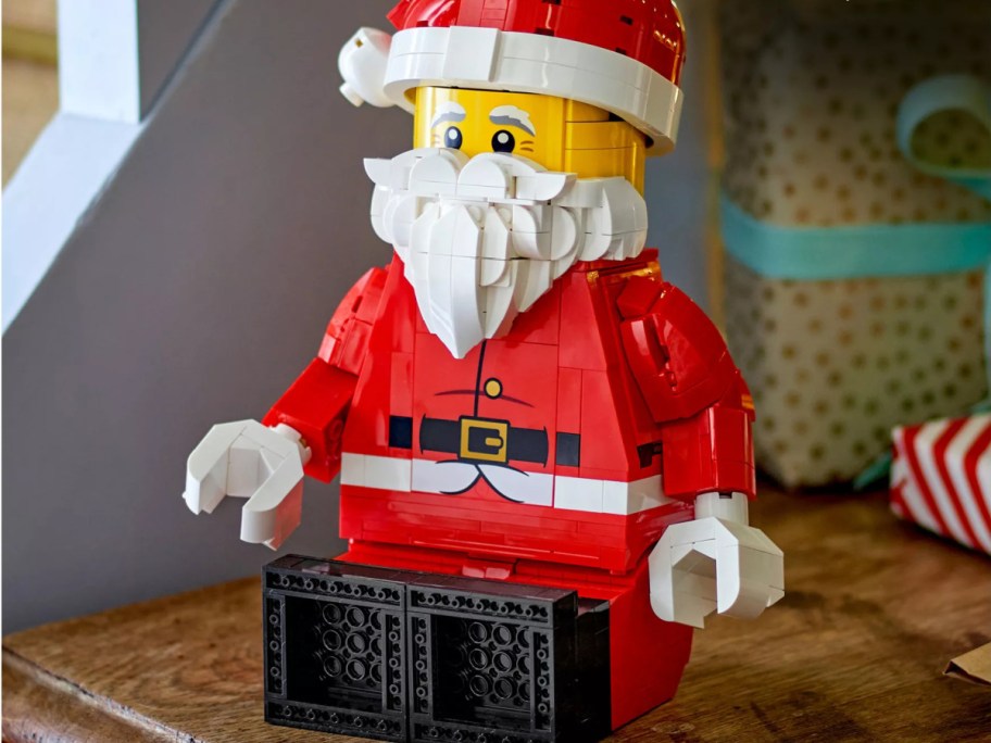 A large, brick-built LEGO Santa Claus figure wearing a red suit and white beard, sitting on a wooden surface.