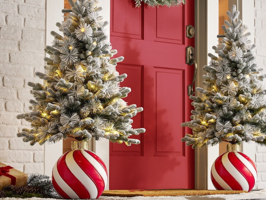 Two pre-lit, flocked miniature Christmas trees displayed in large red-and-white striped ornament bases flanking a bright red front door.