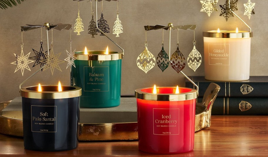 Four scented candles in colored glass jars with gold lids, topped with spinning metal carousel ornaments featuring stars, trees, and holiday motifs.