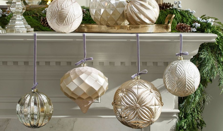 A collection of ornate, shatterproof champagne and silver Christmas ornaments hanging by velvet ribbons from a fireplace mantel decorated with evergreen garland.
