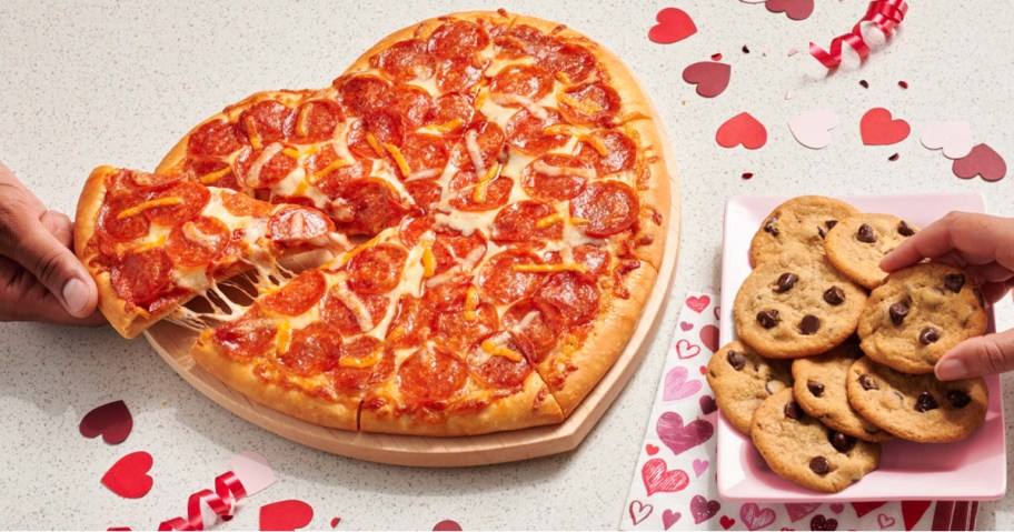 Heart shaped pepperoni pizza on table top next to chocolate chip cookies