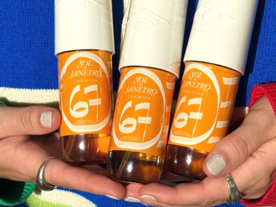 A person holding 3 bottles of yellowish ornage Sol de Janeiro Cheirosa 67 Perfume Mist