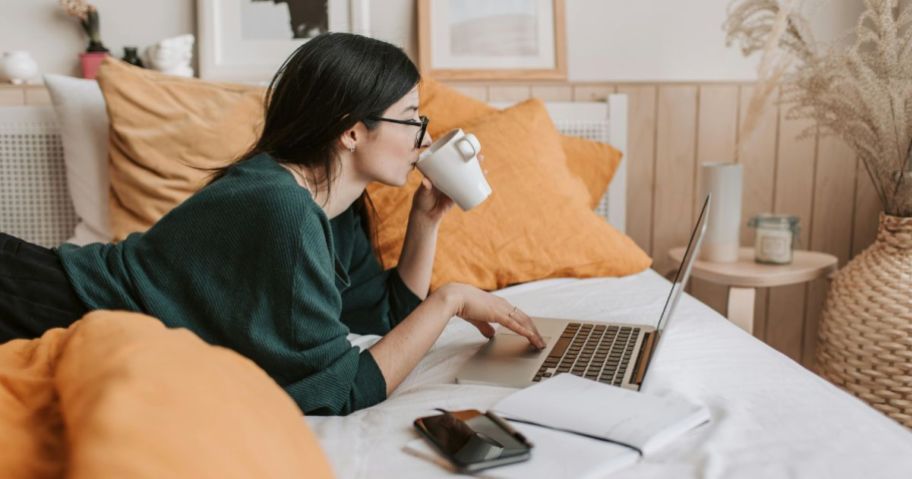 Woman laying on a bed drinking coffee and using a laptop