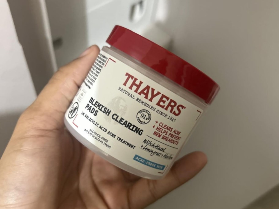 A hand holding a container of Thayers acne pads.