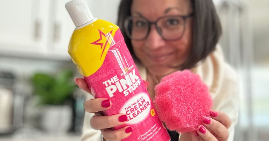 woman holding up a bottle of The Pink Stuff Miracle Cream Cleaner and a pink sponge