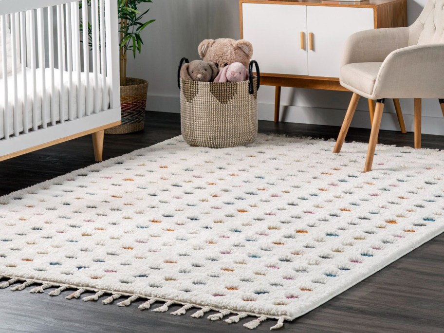 Cozy nursery with a white patterned rug, wicker basket of plush toys, crib, chair, and cabinet against a gray wall.