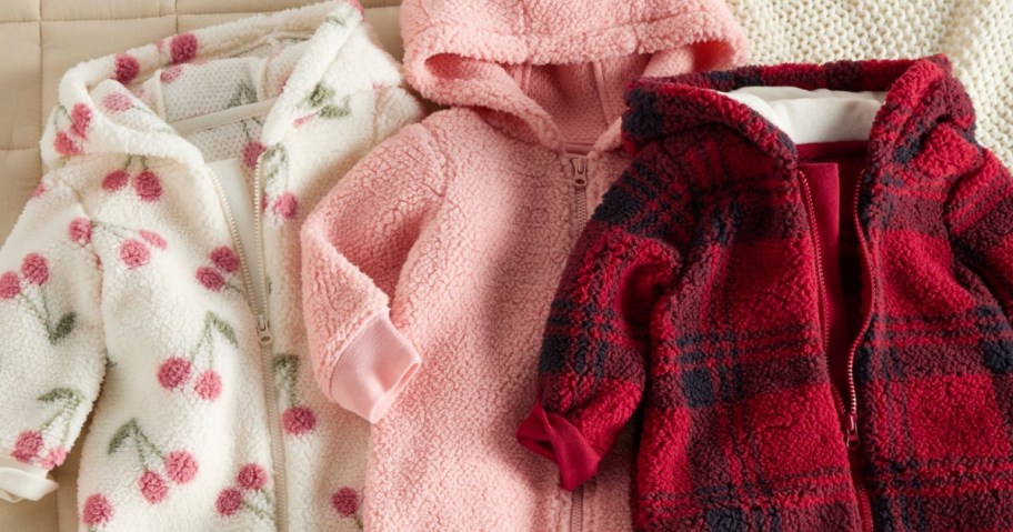Three cozy baby hooded jackets lie side by side on a soft surface: one with pink cherries on white, one solid pink, and one red with black plaid.