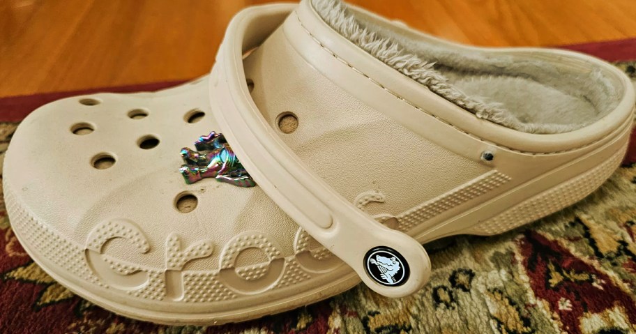 A single beige Croc shoe with fuzzy lining sits on a patterned rug. It features a shiny frog charm on top and numerous round ventilation holes.