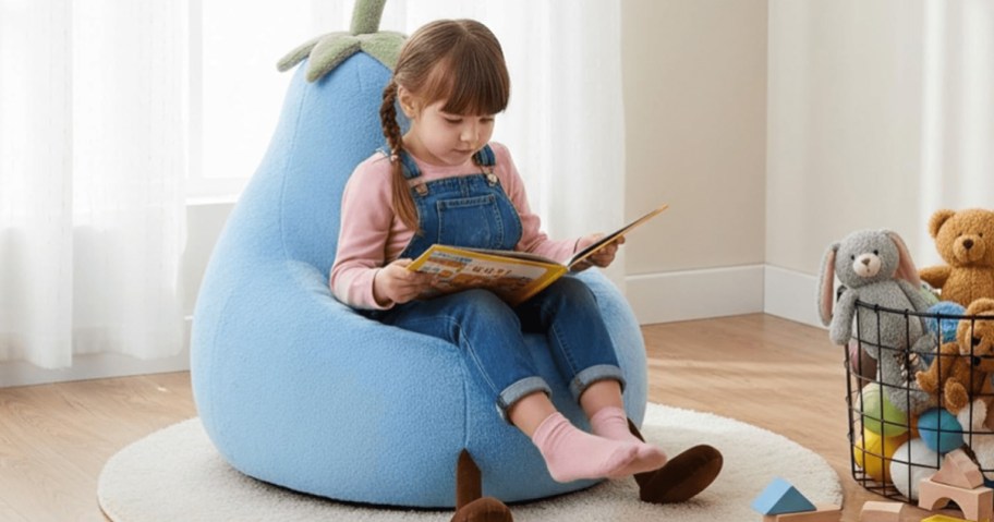 A young girl sitting in a blue eggplant-shaped bean bag chair, reading a book. She is wearing overalls and has light brown hair, with stuffed animals nearby.