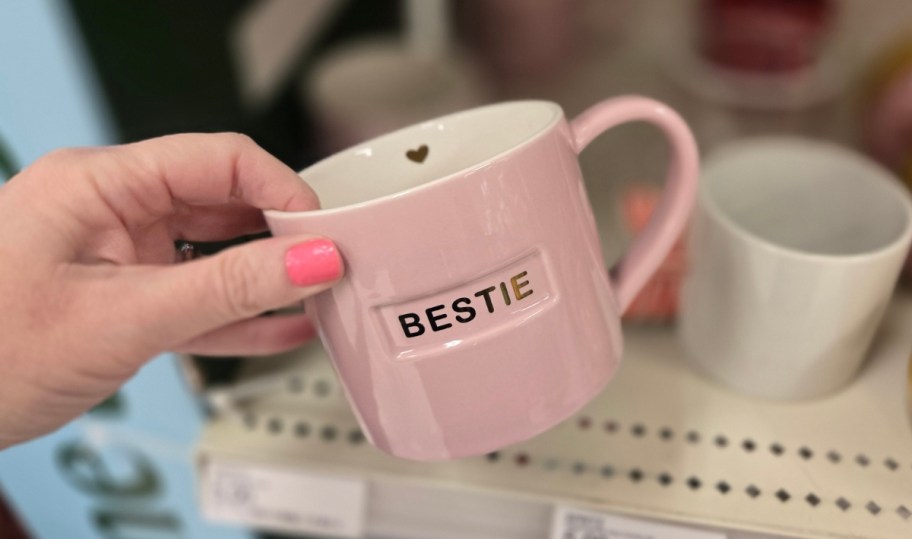 a womans hand holding a pink mug with the word bestie painted on it in gold