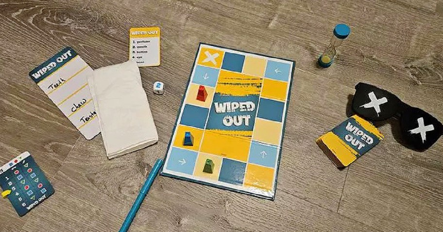 Board game setup on a wooden floor, featuring a blue and yellow board, game cards, dice, timer, pen, notepad, and eye mask with white X's.