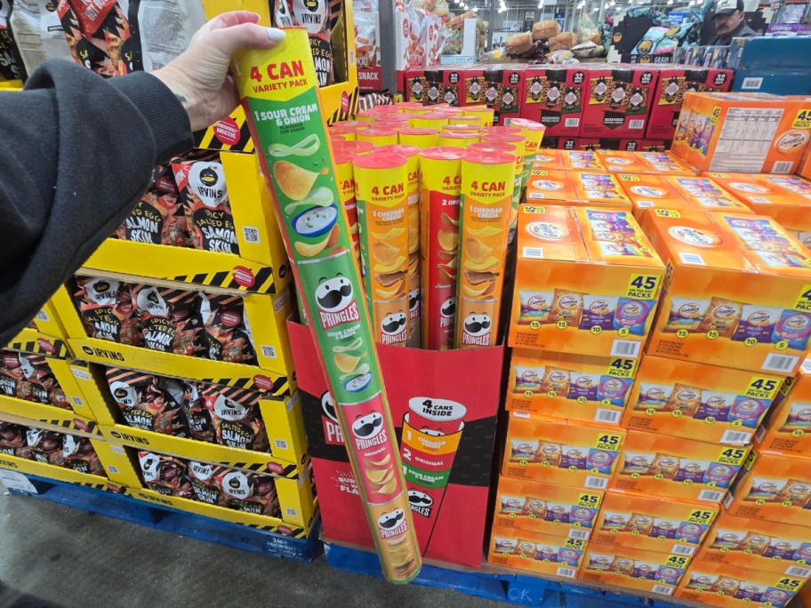 A hand holds a tall Pringles variety pack with four canisters, featuring flavors like sour cream and onion. They stand amidst colorful snack displays in a store.