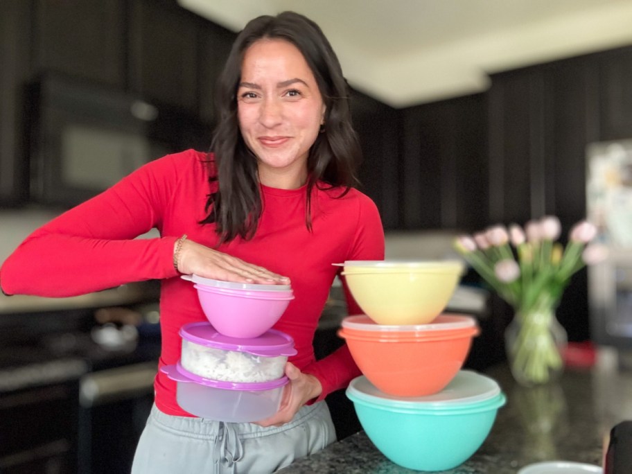 A smiling woman in a red shirt stands in a kitchen, holding stacked colorful bowls and containers. Tulips are on a counter.