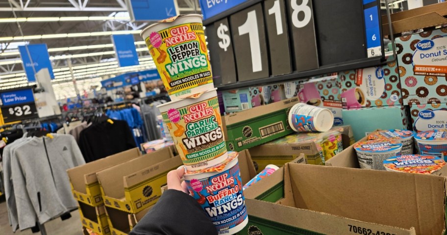 hand holding 3 Nissin Cup Noodles Wing Flavors in front of a display of them at Walmart