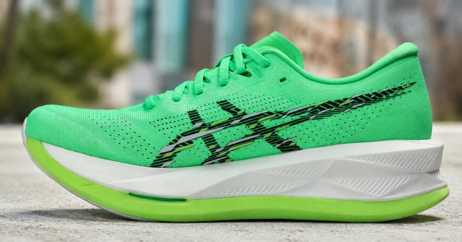A bright green ASICS running shoe with black accents and a thick white midsole sitting on a concrete surface outdoors.