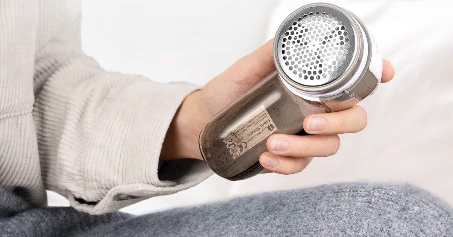 A person's hand holding a fabric shaver, with a perforated metal head, over a sweater.