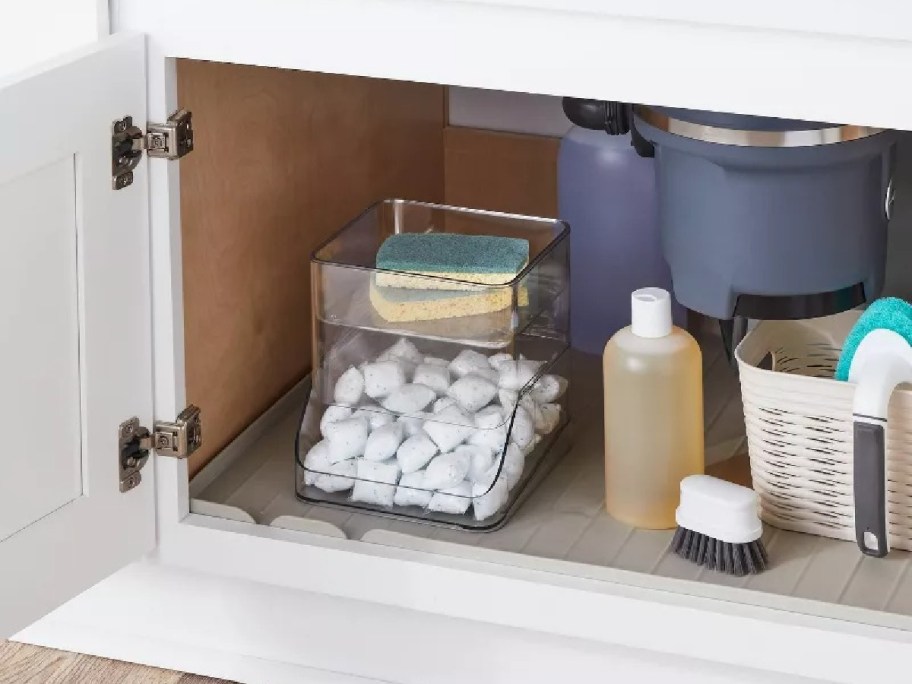 Open kitchen cabinet with cleaning supplies. Includes a container of white pods, two sponges, a dish soap bottle, a brush, and a basket on a liner.