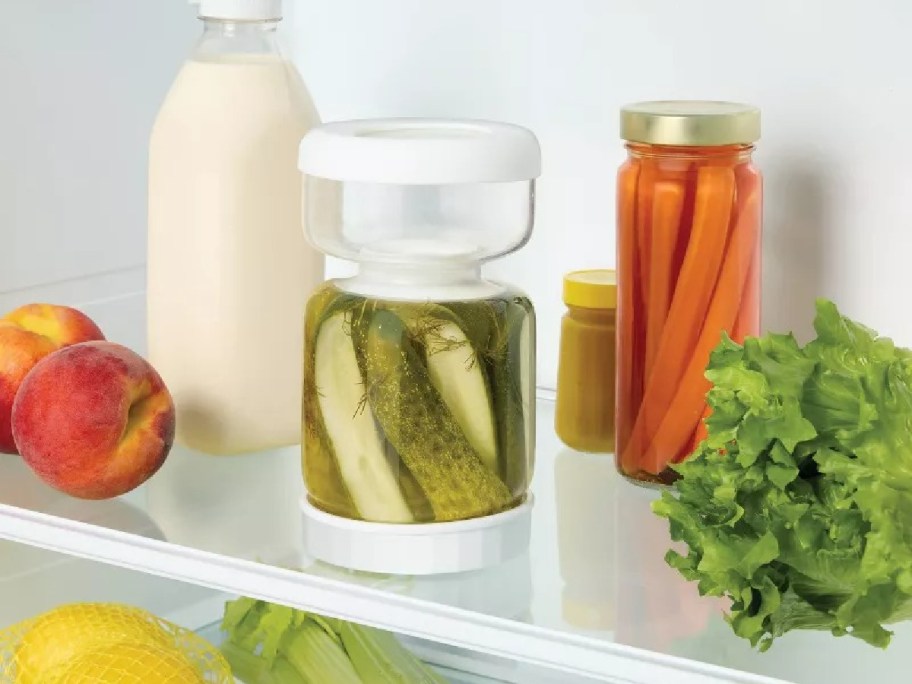 Fridge shelf with a clear container of pickles, a jar of carrot sticks, a lettuce head, two peaches, a milk bottle, and a small mustard jar.