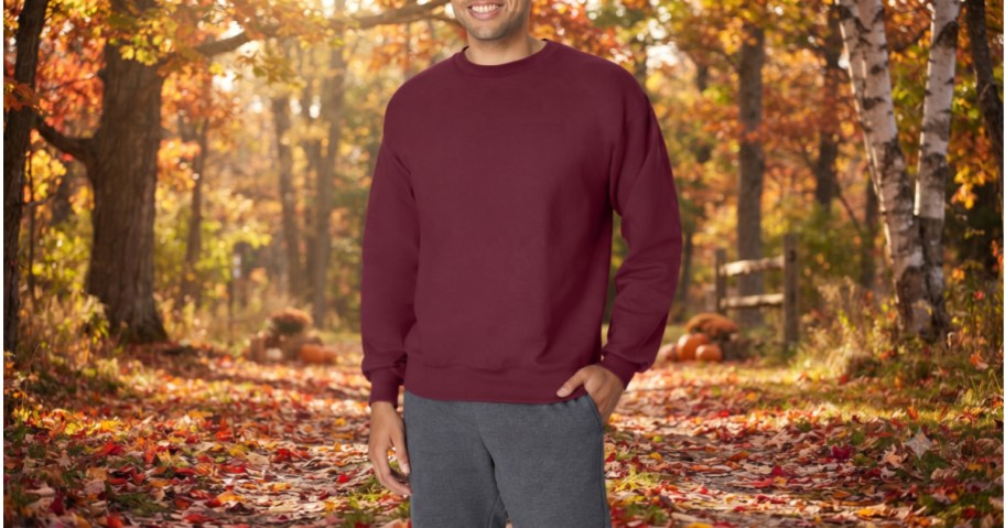 A man wearing a maroon crewneck sweatshirt and grey sweatpants while standing on an autumnal trail covered in fallen leaves.