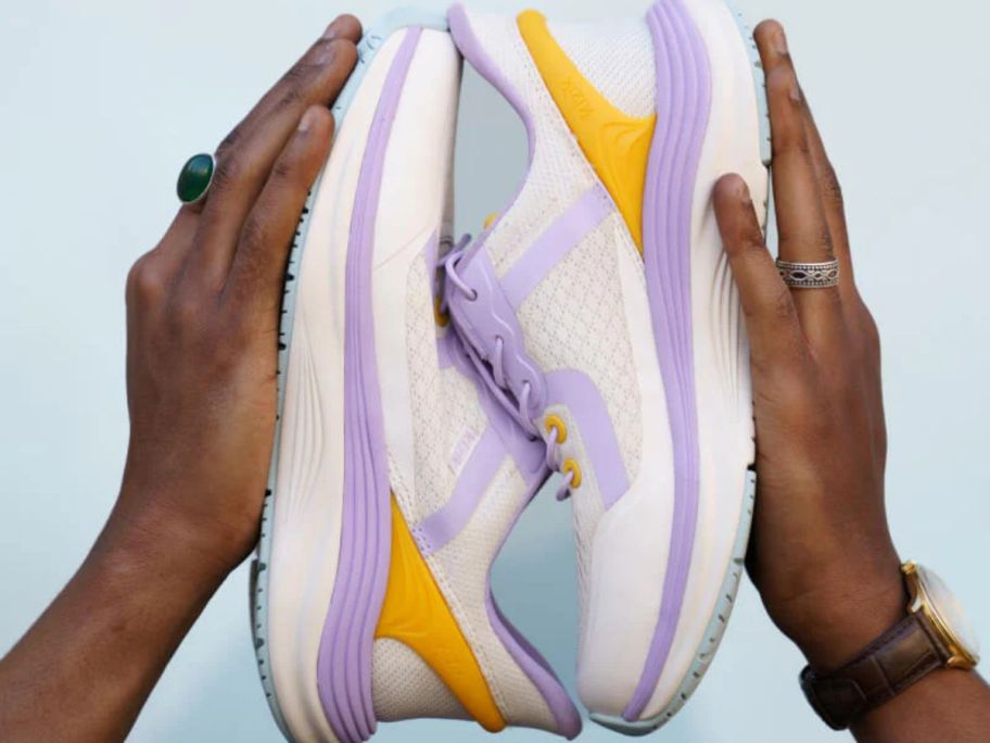 A pair of white sneakers with purple and yellow embellishments being held in between two hands