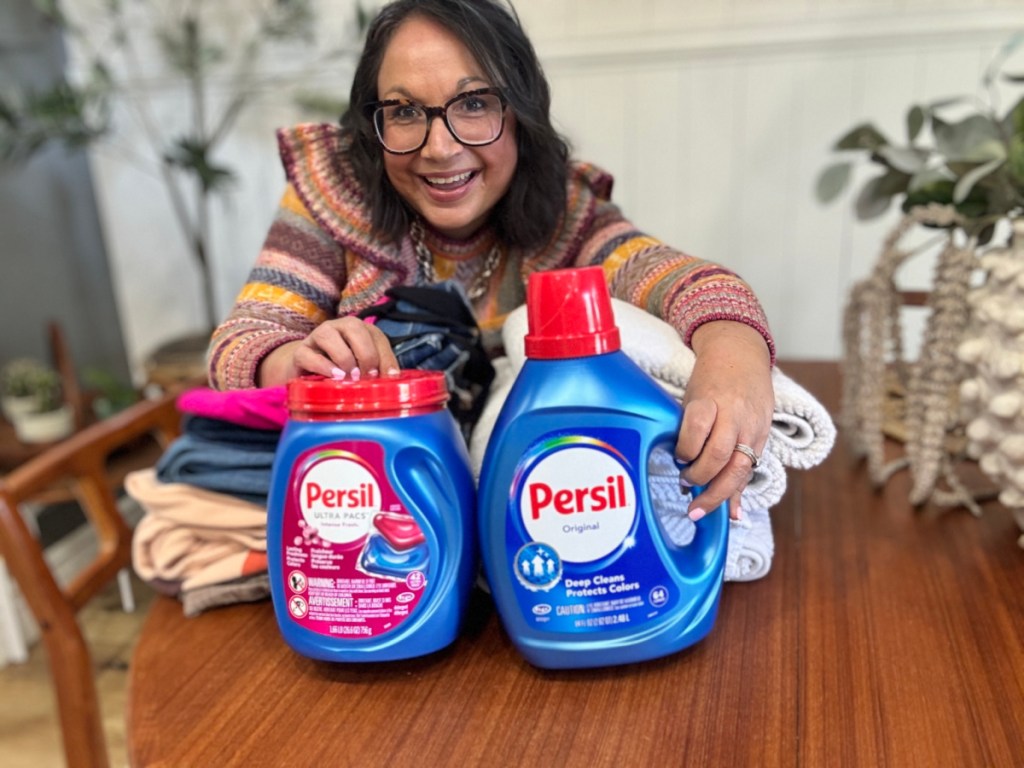 Woman holding two bottle bottles of persil laundry detergent while leaning over table and load of laundry
