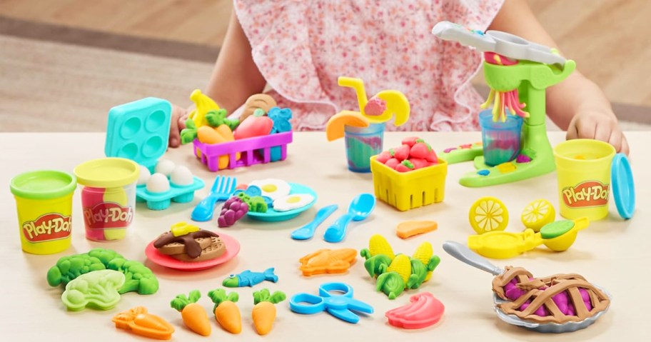 girl sitting at table with Play-Doh Farmer's Market Kitchen Playset laid out in front of her