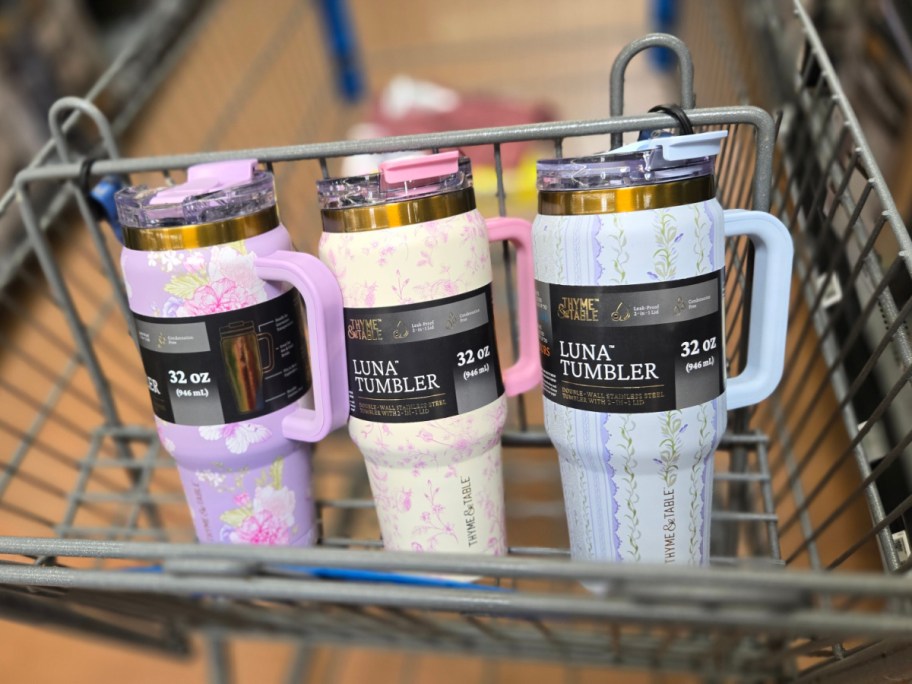 Three large Thyme & Table travel tumblers in pastel colors sit in a shopping cart. The left tumbler is purple with pink flowers, the middle is cream with a pink floral pattern, and the right is light blue with a green vine design.